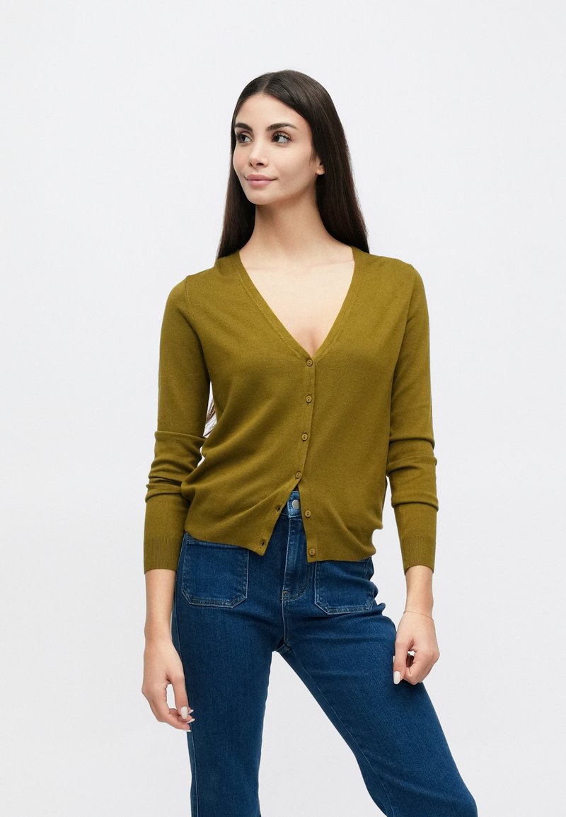 Cardigan vert olive avec col en V, boutonnage sur le devant et manches longues. Assorti à un jean taille haute bleu foncé, texture lisse.