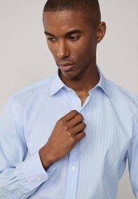 Camicia a righe azzurre chiaro in cotone. Presenta un colletto classico e apertura con bottoni. Maniche lunghe con polsini abbottonati.