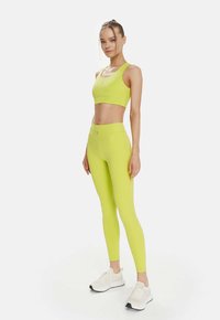 Set de sport format dintr-un sutien și colanți de culoare galben neon, cu o textură netedă. Prezintă un design ajustat și detalii de coasere, împreună cu teniși albi.