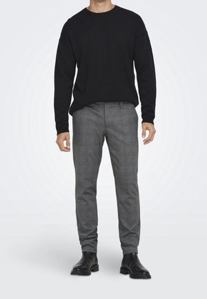Man draagt een zwart trui met lange mouwen, grijze slim-fit broek met patroon en zwarte leren laarzen, staand tegen een witte achtergrond.