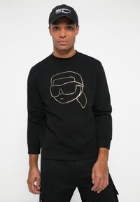 Uomo che indossa un felpa nera con una grafica minimale dorata di un volto e un cappellino nero con testo bianco su sfondo bianco.