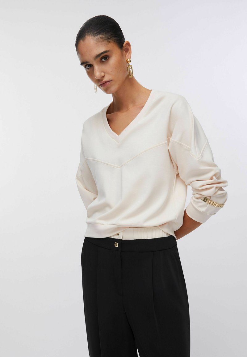 Sweat-shirt crème à col en V avec épaules tombantes, poignets côtelés, détails cousus ; associé à un pantalon noir avec un bouton doré.