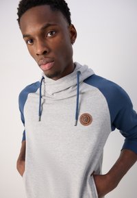 Sudadera gris con mangas azules, un cuello tipo cowl y cordones. Cuenta con un logotipo bordado marrón en el pecho y bolsillos laterales.