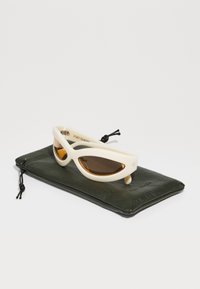 Kattöga solglasögon med en krämfärgad plastram och bruna linser, vilande på en grön läderpouch med dragsko.