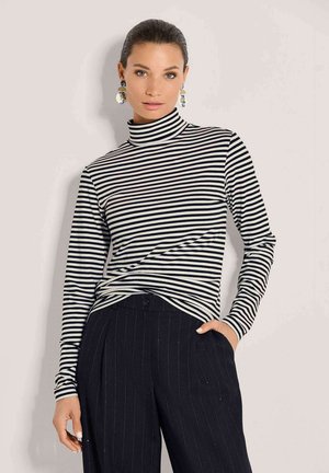 Femme aux cheveux foncés vêtue d’un col roulé à rayures noires et blanches à manches longues et d’un pantalon noir à fines rayures, portant de grandes boucles d’oreilles, debout devant un fond uni.