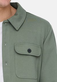 Nahaufnahme einer grünen Jacke mit einer geknöpften Brusttasche, getragen über einem weißen Hemd, zeigt Kragen- und obere Schulternähte.