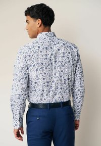 Cămașă albă cu mâneci lungi, cu un model floral albastru, asortată cu pantaloni albaștri. Cămașa are un guler standard și un design cu nasturi.