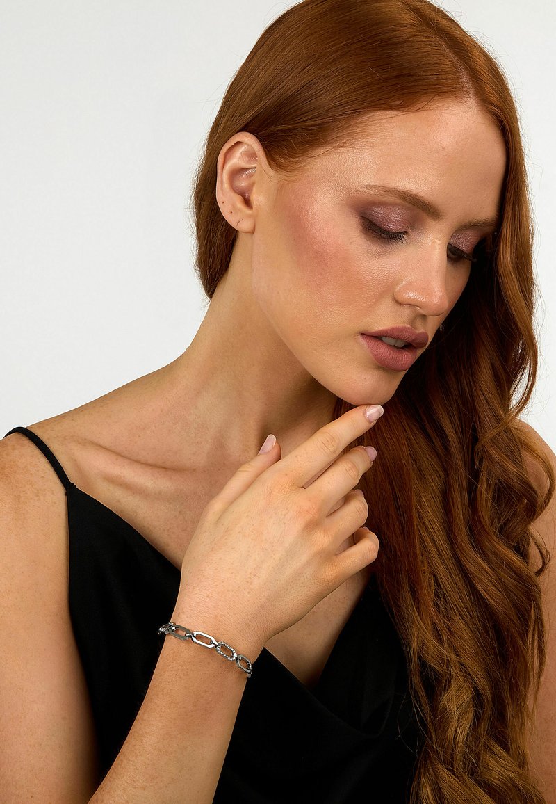 Bracciale in argento con legami ovali e circolari alternati, indossato su una mano con pelle chiara. Texture liscia contro abbigliamento nero.