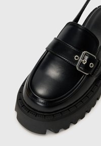 Black leather platform shoe with a rounded toe, wide strap and silver buckle detail, featuring a textured black rubber sole.