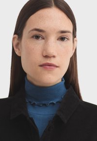 Čierna kordurová košeľa s golierom, vrstvená nad modrým rebrovaným rolákom so strapcovými okrajmi. Hladká textúra a priliehavý dizajn.