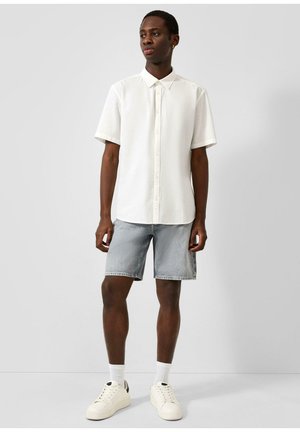 Jeune homme debout portant une chemise blanche à manches courtes boutonnée, un short en denim clair, des chaussettes blanches et des baskets blanches sur un fond uni.