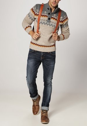 Pletený svetr se vzory v šedé, krémové a oranžové barvě, dvěma knoflíky a koženými popruhy; kombinovaný s modrými džínami a hnědými šněrovacími botami.