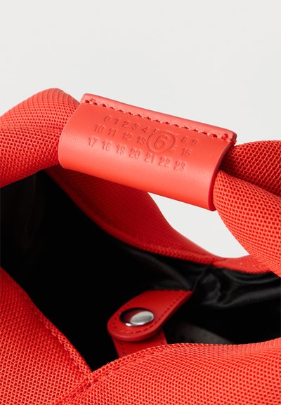 Κοντινή λήψη της λαβής κόκκινης υφασμάτινης τσάντας με ραμμένη δερμάτινη ετικέτα όπου είναι χαραγμένοι αριθμοί από το 0 έως το 23, με τον αριθμό 6 μέσα σε κύκλο να ξεχωρίζει.