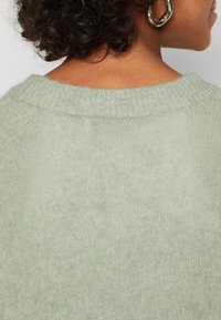 Pletený svetr z měkké, světle zelené látky; má žebrovaný výstřih a texturovaný povrch, což zdůrazňuje jeho útulný a pohodlný vzhled.