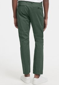 Mörkgröna byxor med rak passform, fram- och bakfickor samt svarta knapppdekorationer. Gjorda av texturerad tyg. Bärs med vita sneakers.