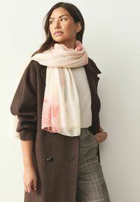 Gaisrs, ar rakstu rakstains šalles gabals maigi rozā un baltās toņos, apsiets ap kaklu, kombinēts ar tumšu mēteļu un rūtainām biksēm.