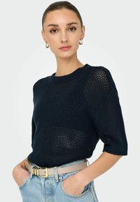 Maglione lavorato a maglia blu navy con vestibilità ampia, maniche corte e un design tessuto a trama aperta. Abbinato a jeans chiari a vita alta e una cintura beige.