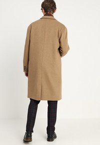 Uomo che indossa un lungo cappotto color cammello, pantaloni a quadri scuri e scarpe nere, in piedi di spalle contro uno sfondo bianco uniforme.