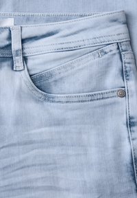 Gros plan de la poche avant d'un jean en denim bleu clair avec des détails de couture, une passants de ceinture et un rivet métallique sur la texture du tissu.
