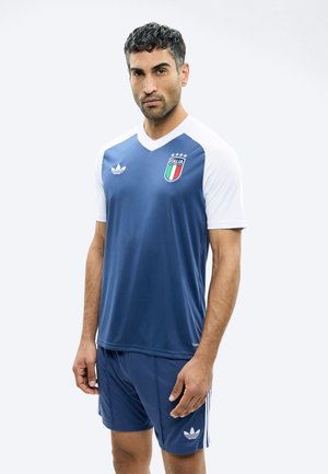 Mann trägt blau-weißes Italia-Fußballtrikot und passende Shorts, steht vor einfachem Hintergrund.