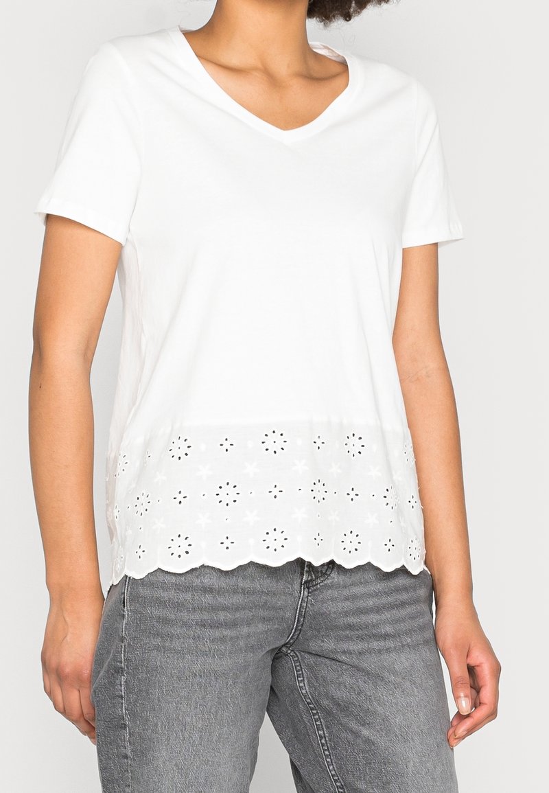 Top bianco a maniche corte con scollo a V e orlo ondulato. La parte inferiore presenta ritagli floreali a occhiello e una texture liscia.