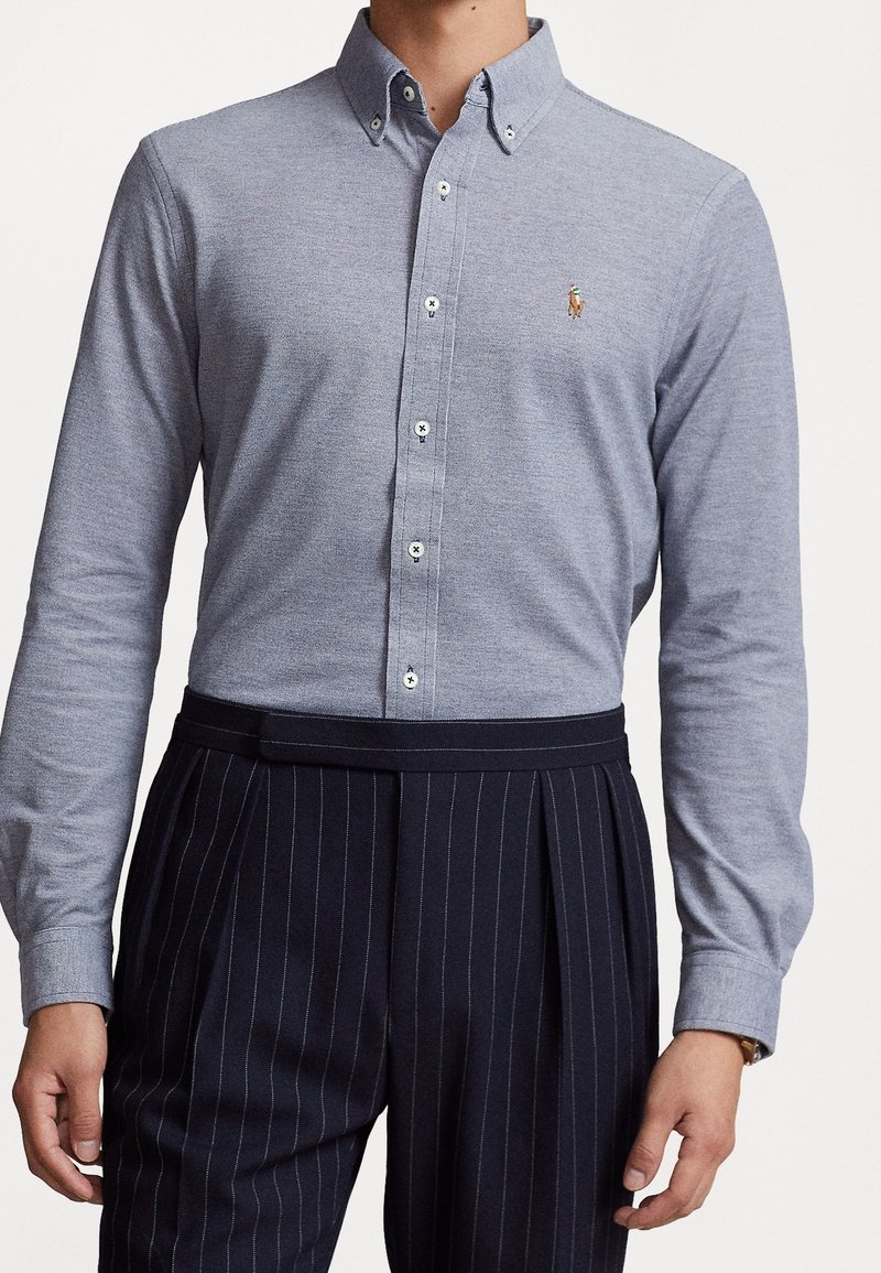 Bílá košile s dlouhým rukávem a knoflíky v světlé šedé barvě z bavlny s malým vyšitým logem, doplněná námořnickými pruhovanými kalhotami.