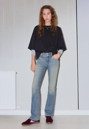 Tânără cu păr ondulat, purtând bluze negre și gri suprapuse, blugi albaștri decolorați și adidași bordo, stând în picioare lângă un perete simplu.