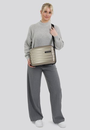 Zilveren harde tas met verticale ribbels, zwart logo, verstelbare schouderband en een rechthoekige vorm, vastgehouden door een persoon in een grijze outfit.