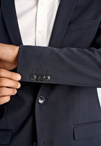 Granatowa marynarka o fakturowanym wykończeniu, z czterema czarnymi guzikami na mankiecie oraz dopasowanym fasonem. Pod spodem widoczna biała koszula.