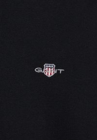 Pull en tricot noir avec un logo GANT brodé en blanc et rouge avec un design en forme de bouclier. Texture lisse et style minimaliste.
