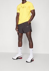 Camisa amarilla de manga corta con estampado "HYROX", pantalones cortos negros, zapatillas blancas con acentos rojos y calcetines blancos de caña corta. Fondo liso.