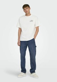 T-shirt en coton blanc avec "After Hours" imprimé en vert, associé à un pantalon cargo en jean bleu et des baskets blanches avec des accents beiges.