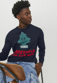 Navy Pullover mit grünem Weihnachtsbaum-Graphic, lächelndem Gesicht und "ROYAL PINE"-Etikett; roter Text mit "CHRISTMAS IN THE AIR". Weiche Textur.