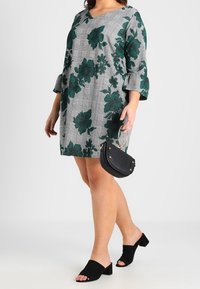 Zielona sukienka w kwiatowy wzór na szarym tle w kratę, z rękawami o długości 3/4, zestawiona z czarnymi, eleganckimi klapkami na obcasie oraz małą czarną torebką.