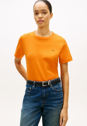 Femeie purtând un tricou portocaliu mulat cu mâneci scurte, băgat în blugi albaștri cu o curea neagră, pe un fundal simplu.