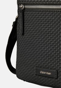 Schwarze gewebte Stofftasche mit Reißverschluss oben, ausgestattet mit einem glatten Lederhenkel und einem kleinen Calvin Klein Logotag. Strukturierte Oberflächendetails.