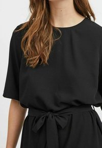 Svart klänning med rund halsringning och korta ärmar. Har ett knytbälte vid midjan, tillverkat av ett mjukt tyg med en behaglig textur.