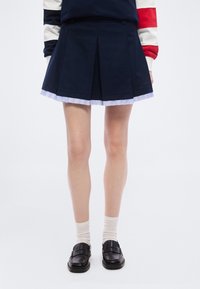 Άτομο που φοράει σκούρα μπλε πλισέ φούστα με λευκή ρίγα στο τελείωμα, λευκές κάλτσες, μαύρα μοκασίνια και πουλόβερ με κόκκινες, λευκές και μπλε μανίκια.