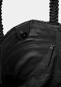 Vnitřek černé tkané tašky s kapsou na zip, zaklapávacím uzávěrem a hladkým povrchem. Rukojeti jsou spletené, což vytváří kontrast s látkou.