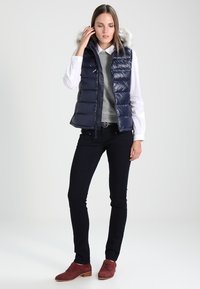 Chaleco acolchado navy con capucha de pelo desmontable, cierre de cremallera y textura acolchada, combinado con un suéter gris y pantalones oscuros.