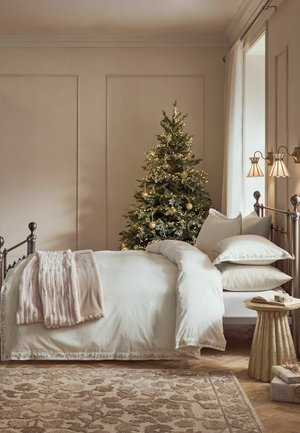 Gemütliches Schlafzimmer mit einem geschmückten Weihnachtsbaum, einem ordentlich gemachten Bett mit weißer Bettwäsche, einer weichen Decke, einem runden Beistelltisch und Geschenkboxen auf einem gemusterten Teppich.