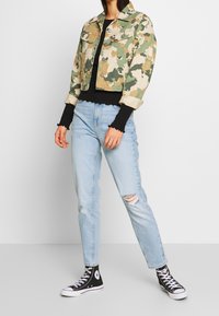 Kamelmönstrad kort denimjacka i beige och grön, kombinerad med en svart långärmad topp och ljusblå slitna jeans. Svarta högsnekers.