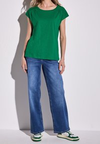 Woman wearing green short-sleeve top, blue wide-leg jeans, and green and white sneakers, standing against a plain light background.