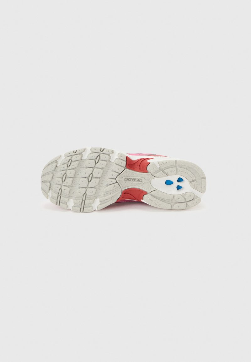 Semelle de chaussure de course avec semelle blanche, accents rouges, éléments d'amorti bleus et logo "new balance" sur un fond clair uni.