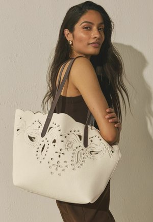 Ung kvinde med langt mørkt hår holder en hvid mulepose med paisley-udskæringsmønstre og mørke skulderstropper.