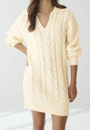 Femme portant une robe-pull crème en maille torsadée avec manches retroussées et col en V, debout devant un fond uni.
