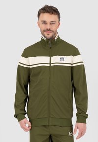 Groene sportjasje met rits, witte horizontale strepen, hoge kraag en zijzakken. Bevat een logo op de borst en geribbelde manchetten.