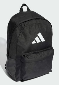 Sac à dos en tissu noir avec une forme arrondie, une poche à l'avant et une poignée supérieure. Présente un grand logo Adidas blanc sur le devant.