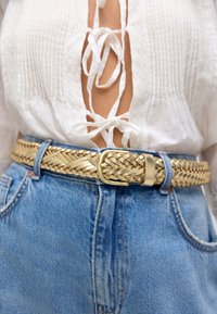 Cinturón trenzado tejido en color dorado con diseño texturizado; usado sobre shorts de mezclilla azul de tiro alto; hebilla de metal plateado; detalle de un conjunto casual.