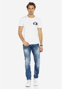 Witte katoenen t-shirt met een zwart bedrukt logo op de borstzak, gecombineerd met blauwe vervaagde slim-fit jeans en witte sneakers.